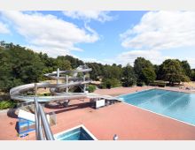 Ein Freibad mit einem großen rechteckigen Schwimmbecken und einer langen, gewundenen Wasserrutsche aus Metall. Im Hintergrund sind grüne Bäume und ein teilweise wolkiger Himmel zu sehen.