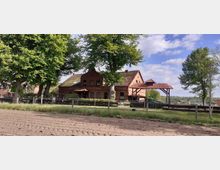 Backsteingebäude im ländlichen Stil mit rotem Ziegeldach, umgeben von Bäumen und eingezäumtem Grünbereich. Im Vordergrund ist ein gepflügtes Feld zu sehen, im Hintergrund erstreckt sich eine bewaldete Landschaft unter blauem Himmel.