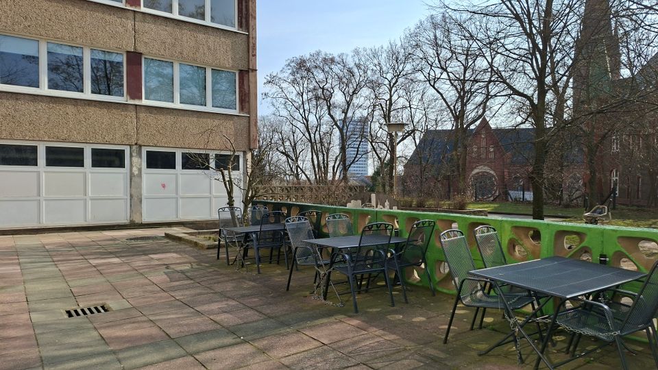 Außenbereich mit gepflasterter Terrasse neben einem mehrstöckigen Gebäude mit großen Fensterreihen und mehreren weißen Garagentoren. Entlang einer niedrigen grün gestrichenen Betonbrüstung stehen Metalltische und -stühle; im Hintergrund sind kahle Bäume, eine rote Backsteinkirche und weitere Gebäude zu sehen.