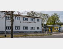 Gebäudeansicht des Werner-Seelenbinder-Stadions in Luckenwalde. Zu sehen ist ein zweistöckiges, schlichtes weißes Gebäude mit Fenstern und aufgebrachter Beschriftung, daneben ein gelber Pavillon mit Eingangsbereich und Sportplatz im Hintergrund, umgeben von Bäumen.