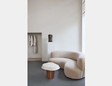 Heller, minimalistischer Innenraum mit einer beigefarbenen, geschwungenen Polstercouch, einem kleinen weißen Beistelltisch mit Holzbasis, einer Büste auf einem Sockel und einer Nische mit hängenden, beigen Kleidungsstücken. Die Wände sind schlicht weiß, der Boden grau.