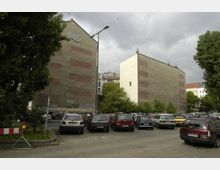 Offener Parkplatz in einer städtischen Umgebung, umgeben von mit Graffiti versehenen Hausfassaden und Bäumen. Im Hintergrund sind Wohngebäude und ein bewölkter Himmel sichtbar.