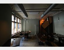 Innenraum eines modernen Restaurants oder Cafés mit grauen Wänden, Betonboden und freiliegenden Lüftungsrohren an der Decke. Links stehen Tische an großen Fenstern mit leichten Vorhängen, rechts weitere Sitzplätze neben einer geometrischen Holzstruktur und hängenden Lampen.