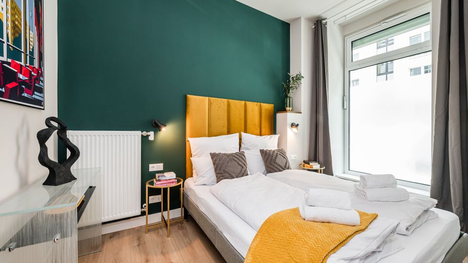 Modernes Schlafzimmer mit zwei Einzelbetten, senfgelbem gepolstertem Kopfteil vor dunkelgrüner Akzentwand und Holzfußboden. Rechts ein großes Fenster mit grauen Vorhängen; daneben kleine Nachttische mit Lampen, links eine Kommode und ein Wandbild.