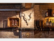 Backsteinwand mit rustikalem Dekor, darunter Cowboy-Stiefel auf einem Regal und einer Holzkiste, ein montierter Antilopenschädel sowie eine Auswahl von Flaschen mit Spirituosen auf einem weiteren Regal. Ein schwarzes Rohr verläuft horizontal entlang der Wand.