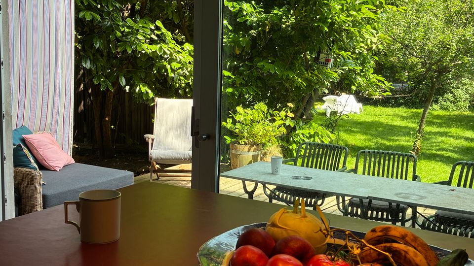 Blick aus einem Essbereich durch eine große Glastür auf eine Terrasse mit langem Tisch und Stühlen sowie einen grünen Garten mit Bäumen und Rasen. Im Vordergrund steht auf dem Tisch eine Schale mit Obst und Gemüse neben einer Tasse.