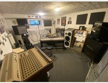 Ein Proberaum mit Musikequipment, darunter ein Mischpult im Vordergrund, ein Keyboard, Schlagzeug, Lautsprecher und ein Mikrofon. Der Raum hat weiße Wände mit Schaumstoffplatten zur Schalldämmung und verschiedenen Postern sowie Fotos zur Dekoration.