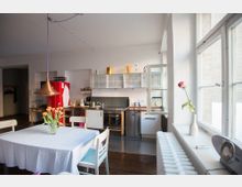 Helle Küche mit Esstisch, Kupferlampe und roten Tulpen in einer Vase. Im Hintergrund eine moderne Küchenzeile mit weißem Schrank, Edelstahlgeräten und einem roten Kühlschrank, daneben ein geöffnetes Fenster mit Blick nach draußen.