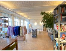 Ein heller, offener Raum mit Holzfußboden, hohen Decken und mehreren großen Fenstern, die viel Tageslicht hereinlassen. Zu sehen sind Regalwände mit Büchern, Kleiderständer mit bunter Kleidung, ein großer Baum im Topf sowie ein Tisch und Büromaterialien im Hintergrund.