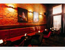 Innenraum einer Bar oder Lounge mit freiliegender Backsteinwand, gerahmten Bildern und warmen Wandleuchten über einer langen, gepolsterten roten Sitzbank. Davor stehen kleine runde Tische mit Tischlampen und dunkle Stühle; rechts fällt Licht durch ein großes Fenster mit Vorhang.