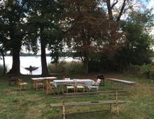 Wiese mit Tischen und Stühlen, die für eine Veranstaltung aufgestellt sind, im Vordergrund eine Holzbank; im Hintergrund Bäume, ein See und eine Hängematte zwischen zwei Bäumen.
