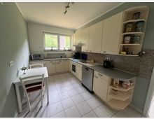 Helle Wohnküche mit L-förmiger Einbauküche in cremefarbenen Schränken, grauer Arbeitsplatte und Fliesenboden, mit Herd, Spüle und Geschirrspüler. Ein großes Fenster zur grünen Außenansicht sowie ein kleiner Esstisch mit zwei Stühlen stehen an der linken Wand.