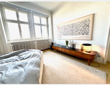Helles Schlafzimmer mit großen Sprossenfenstern und Heizkörper darunter, einem Bett im Vordergrund und einem langen, niedrigen Sideboard an der Wand. Über dem Sideboard hängt ein großes gerahmtes Wandbild; auf dem Möbel stehen Bücher, Glasflaschen und eine Tischlampe auf hellem Teppichboden.