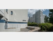 Mehrstöckige Wohngebäude aus Beton in einem urbanen Wohnviertel, umgeben von Büschen, Bäumen und gepflasterten Wegen; im Vordergrund ein kleiner Eingangsbereich mit Rampe und Treppe, eingefasst durch Geländer.