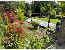 Garten mit üppiger Vegetation, einer Mischung aus roten und grünen Sträuchern sowie kleinen Bäumen. Im Hintergrund ein gepflegter Rasen mit Trampolin, einem Pool mit Abdeckung und einer Löwenstatue aus Stein.