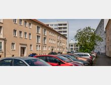 Straßenszene mit geparkten Autos vor einem beige gestrichenen Wohngebäude mit mehreren Fenstern und einem roten Ziegeldach. Im Hintergrund ist ein moderneres, mehrstöckiges Gebäude sowie ein Baum auf der rechten Seite zu sehen.