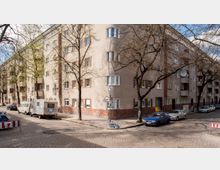 Wohngebäude an einer Straßenecke mit hellbeiger Fassade, roten Backsteinelementen im unteren Bereich und mehreren Fenstern. Am Straßenrand sind geparkte Autos und Wohnwagen zu sehen, umgeben von kahlen Bäumen auf einer Kopfsteinpflasterstraße.