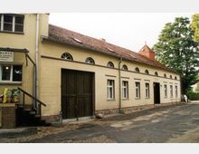 Ein langgestrecktes, gelb verputztes Gebäude mit einem roten Ziegeldach und halbrunden Fenstern unter der Dachkante. Links befindet sich ein großes, dunkles Holztor, daneben mehrere Fenster und ein Eingang; rechts ist ein kleiner Turm sichtbar.