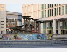 Springbrunnen auf einem Platz in einer urbanen Umgebung, mit einer runden, kunstvoll bemalten Umrandung und mehreren säulenartigen Skulpturen aus Metall. Im Hintergrund sind moderne Gebäude mit großen Glasfassaden sowie ein Eingangsbereich mit goldenen Türen zu sehen.