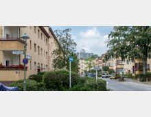 Wohnsiedlung mit mehrstöckigen gelb verputzten Gebäuden, Balkonen und roten Fensterrahmen, umgeben von grünen Büschen und Bäumen. Im Vordergrund Straßenschilder, parkende Autos und ein Gehweg auf einer Kopfsteinpflasterstraße.