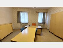 Ein schlicht eingerichteter Büroraum mit zwei Holztischen, drei Stühlen und einem grauen Telefon auf dem Tisch. Im Hintergrund befinden sich zwei Fenster mit Gardinen, unter denen Heizkörper angebracht sind, und die Wände sind mit hellem Tapetenmuster bedeckt.