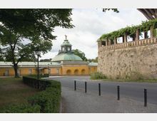 Historisches Gebäude mit gelber Fassade, grüner Kupferkuppel und Uhrturm, umgeben von Bäumen und einer Kopfsteinpflasterstraße. Rechts im Bild eine alte Steinmauer mit Pergola und Begrünung.