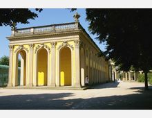 Gelbes, klassizistisches Gebäude mit hohen Rundbogenfenstern, verziert mit Säulen und floralen Reliefs, umgeben von Bäumen und einem offenen Hof mit Kiesboden. Im Hintergrund sind weitere Säulenreihen und Grünflächen zu sehen.