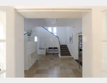 Heller Eingangsbereich eines Gebäudes mit Fliesenboden, einer Treppe mit grauem Teppichbelag in der Mitte, einem weißen Schrank links und zwei Stühlen daneben. An der rechten Wand hängt ein gerahmtes Kunstwerk, und im Hintergrund ist eine Tür zu einem Raum mit Bücherregalen erkennbar.