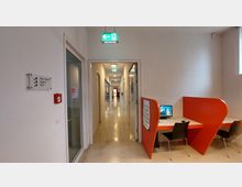 Ein heller Flur in einem modernen Gebäude mit Türen auf beiden Seiten, einem Fluchtwegschild an der Decke und einem Glastürbereich links. Im Vordergrund befindet sich eine Arbeitsecke mit zwei orangefarbenen, abgegrenzten Arbeitsplätzen, einem Computer und Stühlen.