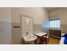 Ein einfacher Raum mit einer schlichten Einrichtung: ein kleines Waschbecken mit Fliesen an der Wand und einem kleinen Spiegel, ein Holztisch mit Hocker sowie ein vergittertes Fenster, das wenig Licht hereinscheinen lässt. Der Raum hat helle Wände und einen Holzboden.