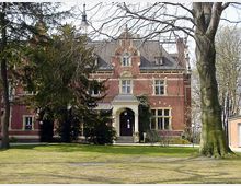 Backsteingebäude im Stil eines historischen Herrenhauses mit verzierten Giebeln, hohen Fenstern und kleiner überdachter Eingangstreppe, umgeben von Bäumen und einer gepflegten Rasenfläche. Im Vordergrund stehen mehrere große Bäume.