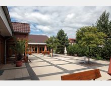 Offener Innenhof mit gepflastertem Boden und modernen Mustern, umgeben von niedrigen Gebäuden mit roten Ziegeldächern. Es gibt Holzbänke, Bäume, Büsche, eine Palme in einem Topf und einen kleinen Außengrill, mit einem bewölkten Himmel im Hintergrund.