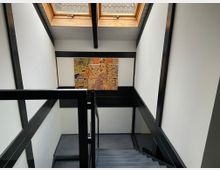 Treppe in einem modern gestalteten Innenraum mit schwarzen Geländern und weißen Wänden, darüber ein Dachfenster mit Holzrahmen. An der Wand hängt ein großes, buntes Gemälde mit einer Frau in einer retroartigen Szene.