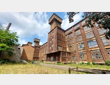 Backstein-Industriegebäude mit mehreren Stockwerken, großen rostfarbenen Fenstern und charakteristischen Wassertürmen im Hintergrund. Im Vordergrund eine verwilderte Wiese mit Gehweg, umgeben von Bäumen und einem verfallenen Zaun.