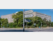 Straßenszene mit Verkehrsschildern, die auf ein Krankenhaus, ein Tiergehege und ein städtisches Feuerwehrmuseum hinweisen, im Vordergrund. Im Hintergrund ein mehrstöckiges, rechteckiges Gebäude mit großen, gleichmäßigen Fenstern, umgeben von Bäumen und Sträuchern.