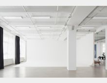 Heller, minimalistischer Studio-Raum mit weißen Wänden und einer Hohlkehle im Hintergrund, großen Fenstern mit dunklen Vorhängen auf der linken Seite und sichtbaren Rohren und Leuchtstofflampen an der Decke. Rechts im Bild befindet sich eine Säule, und im angrenzenden Bereich stehen einige Möbel.