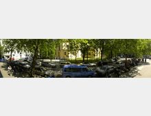 Panoramaaufnahme eines städtischen Platzes mit parkenden Autos und Motorrädern unter Bäumen. Im Hintergrund ist ein gelb gestrichenes Gebäude mit historischer Architektur erkennbar, umgeben von einem Zaun und Grünanlagen.