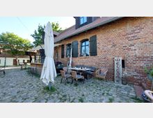 Backsteinbau mit schwarzen Fensterläden und einem Außenbereich, bestehend aus Tischen, Stühlen und einem Sonnenschirm auf einem gepflasterten Hof. Rechts steht ein Holzschild mit der Aufschrift „Welcome“, im Hintergrund befinden sich weitere Gebäude und Bäume.