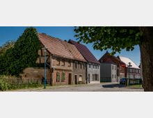 Straßenszene in einer ländlichen Umgebung mit alten, teilweise verfallenen Wohnhäusern aus Ziegel- und Putzbau, flankiert von Bäumen und einem Kopfsteinpflasterweg. Im Hintergrund befinden sich weitere Gebäude, darunter ein moderneres Haus und ein Fachwerkhaus.