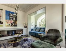 Wohnzimmer mit modernem Design; im Vordergrund grüne Polstermöbel und drei schwarze Tische mit goldenen Details, im Hintergrund ein großes Sofa in Blau vor einem großen, bogenförmigen Fenster. An der Wand befindet sich ein Kamin mit dekorativen Elementen und zwei Kunstbilder.