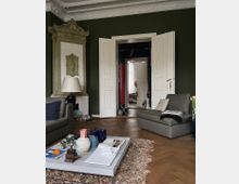 Wohnzimmer mit olivgrünen Wänden, weißen Doppeltüren und reich verziertem Stuck an der Decke. Im Vordergrund stehen ein Couchtisch mit Dekorationsgegenständen, zwei graue Sofas und ein Teppich über einem Parkettboden. Im Hintergrund führt der Blick durch geöffnete Türen in angrenzende Räume.