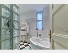 Ein Badezimmer mit hellen Fliesenwänden, einer Eckbadewanne, blickdichten Fenstern und einem Handtuchheizkörper, auf dem Handtücher hängen. Links ist eine halbtransparente Trennwand aus Glasbausteinen zu sehen, der Boden hat ein schwarz-weißes Schachbrettmuster.