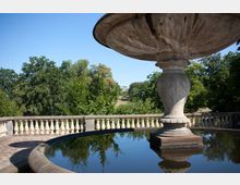 Großer steinerner Brunnen mit einer flachen Schale, die sich in einem kleinen Wasserbecken spiegelt, umgeben von einem Geländer mit Balustern. Im Hintergrund sind grüne Bäume und ein historisches Gebäude in der Ferne erkennbar.