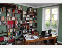 Arbeitszimmer mit grünen Wänden, einem großen Regal voller Bücher und Dokumente sowie einem Holzschreibtisch. Vor dem Fenster, das den Blick in einen grünen Garten gewährt, stehen Computer und Büroartikel auf dem Schreibtisch.