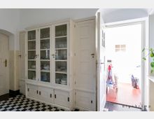 Ein heller Raum mit einem weißen, hohen Schrank mit Glastüren, die Geschirr und Glaswaren zeigen. Der Boden ist mit schwarz-weißen Fliesen gestaltet, und durch eine geöffnete Tür gelangt Licht aus einem angrenzenden Raum mit roten Bodenfliesen hinein.