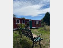 Eine rote Holzhütte mit weißen Fensterrahmen und einer grünen Tür steht in einer ländlichen Umgebung, umgeben von Gras, Wildpflanzen und Blumenkästen. Im Vordergrund befindet sich eine schmiedeeiserne Bank mit einer grünen Holzsitzfläche.