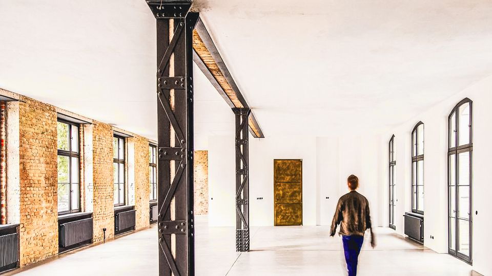 Großer, heller Industrie-Loft-Innenraum mit sichtbaren Stahlträgern und Backsteinwänden entlang der linken Seite. Hohe Fensterreihen und Türen auf beiden Seiten sowie ein glatter Betonboden prägen den offenen Raum.