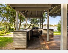 Überdachter Grillplatz aus Holz mit eingebautem Gasgrill und umlaufender Arbeitsfläche auf einer Terrasse. Im Hintergrund liegen Wiese, Bäume und ein Seeufer mit Blick auf Häuser am gegenüberliegenden Ufer.