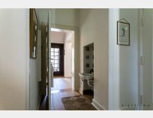 Schmaler Flur in einem Wohnhaus mit weiß gestrichenen Wänden, Holzboden und einer offenen Tür, die zu einer verglasten Eingangstür mit schwarzen Rahmen führt. Auf der rechten Wand befindet sich ein kleines Waschbecken unter einer eingezogenen Nische, und links sind Bilderrahmen aufgehängt.