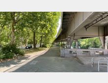 Fußweg unter einer erhöhten Brückenkonstruktion, flankiert von Bäumen und einer Straße auf der linken Seite. Im Hintergrund ist ein mit Pflanzen gesäumter Kanal zu sehen, die Betonpfeiler der Brücke sind mit Graffitis bemalt.